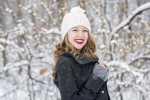 Smiling young woman in a white knit beanie and black coat in a snowy forest setting.