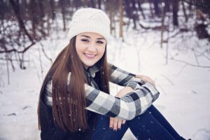 Smiling young woman sits in a snowy forest wearing a white knit beanie and plaid shirt.