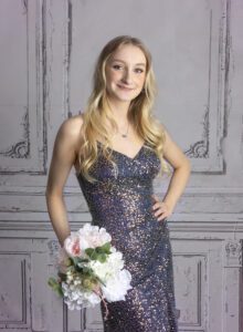 Smiling young woman in a navy sequined dress holds a pink and white bouquet against a light, textured backdrop.