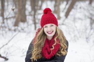 Woman in a red knit hat and scarf smiles in a snowy forest setting.